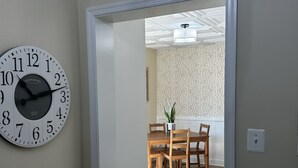 Corredor com relógio fixado na parede, piso de madeira e porta de entrada para uma sala de jantar com mesa e cadeiras.