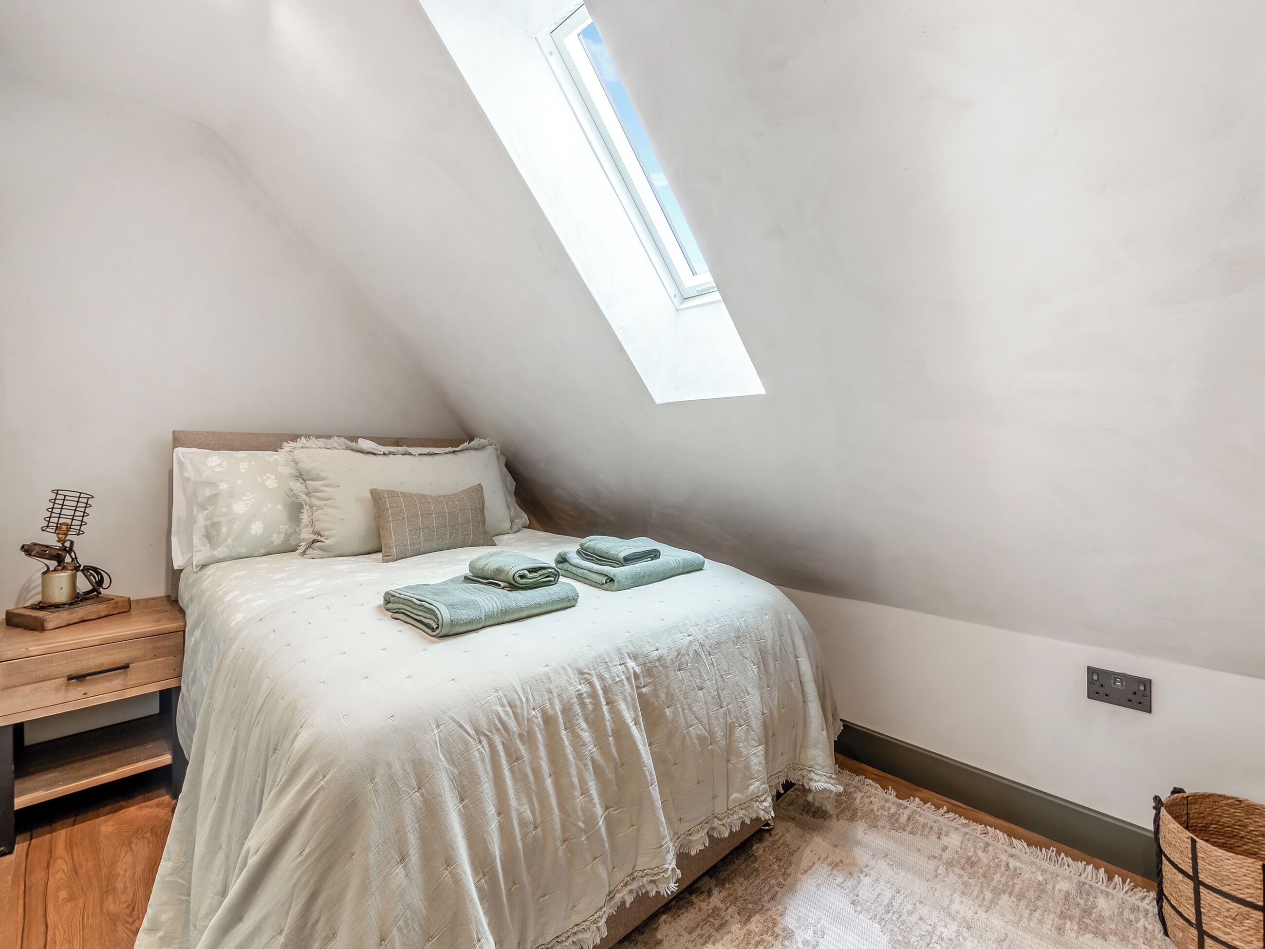 A bedroom with a sloped ceiling, a bed with a white duvet, two pillows, a small bedside table with a lamp, and a window providing natural light.