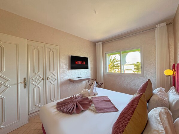 A hotel room with a bed, a TV mounted on the wall, a window with a view of palm trees, and a door with decorative panels. - Luxurious Duplex in a Peaceful Resort (Marrakech)
