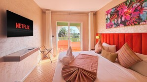 A hotel room with a bed, a TV displaying 'NETFLIX', a colorful floral painting, and a balcony with a view of palm trees. - Luxurious Duplex in a Peaceful Resort (Marrakech)