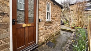Una casa de piedra con puerta de madera, una ventana pequeña y un jardín con plantas y un árbol.