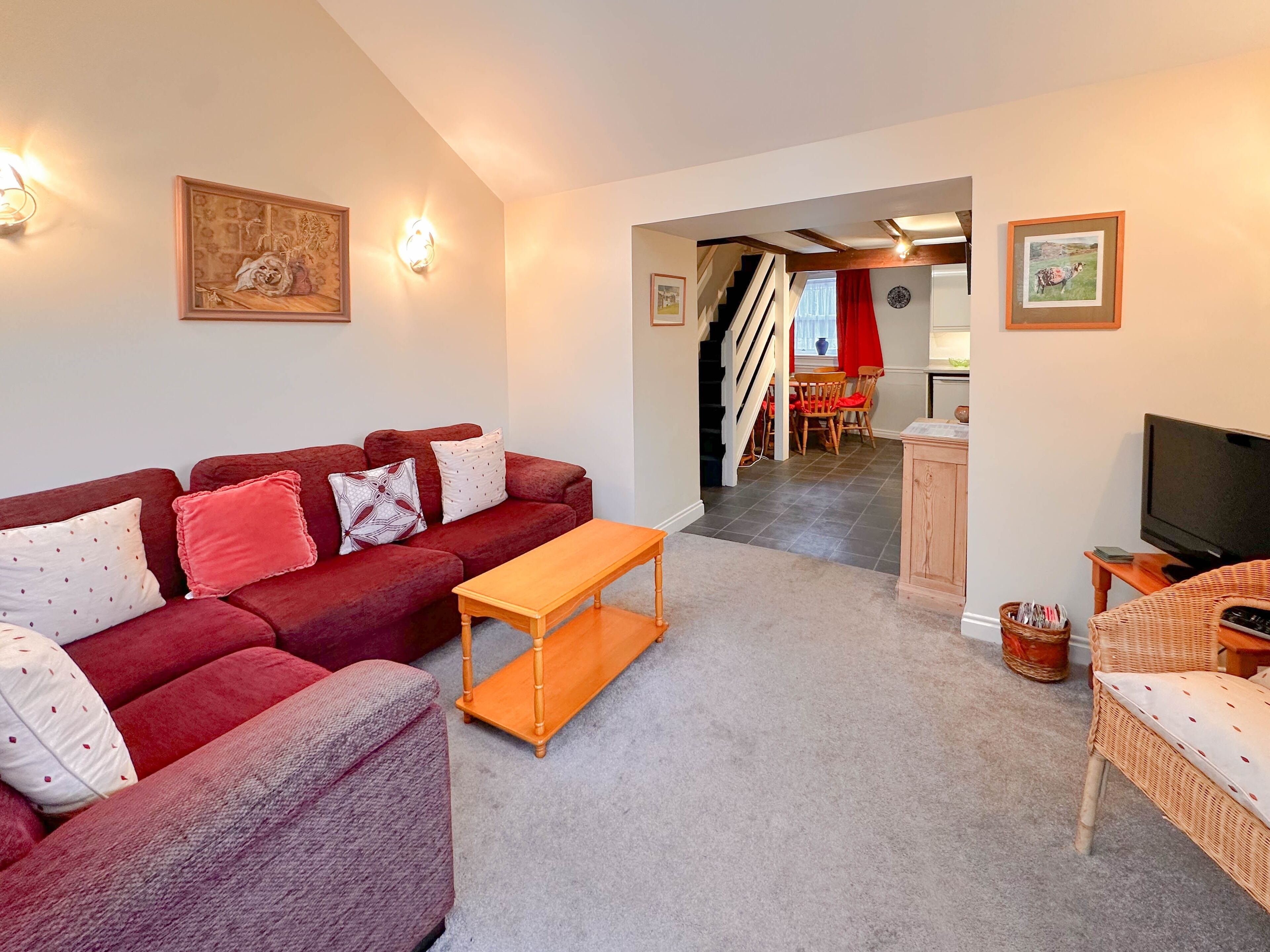 A living room with a red sofa, a wooden coffee table, and a television. There is a staircase leading to a dining area with a red table and chairs.