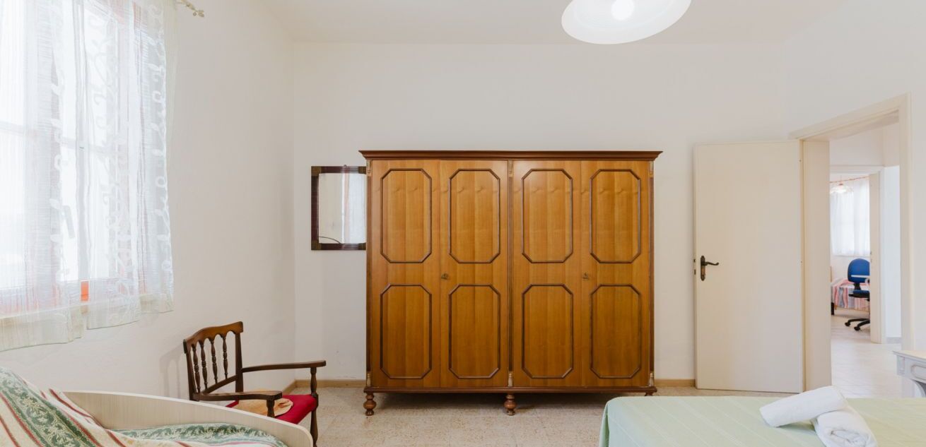 Un dormitori amb un armari de fusta, una cadira, un llit i una finestra amb cortines lleugeres.