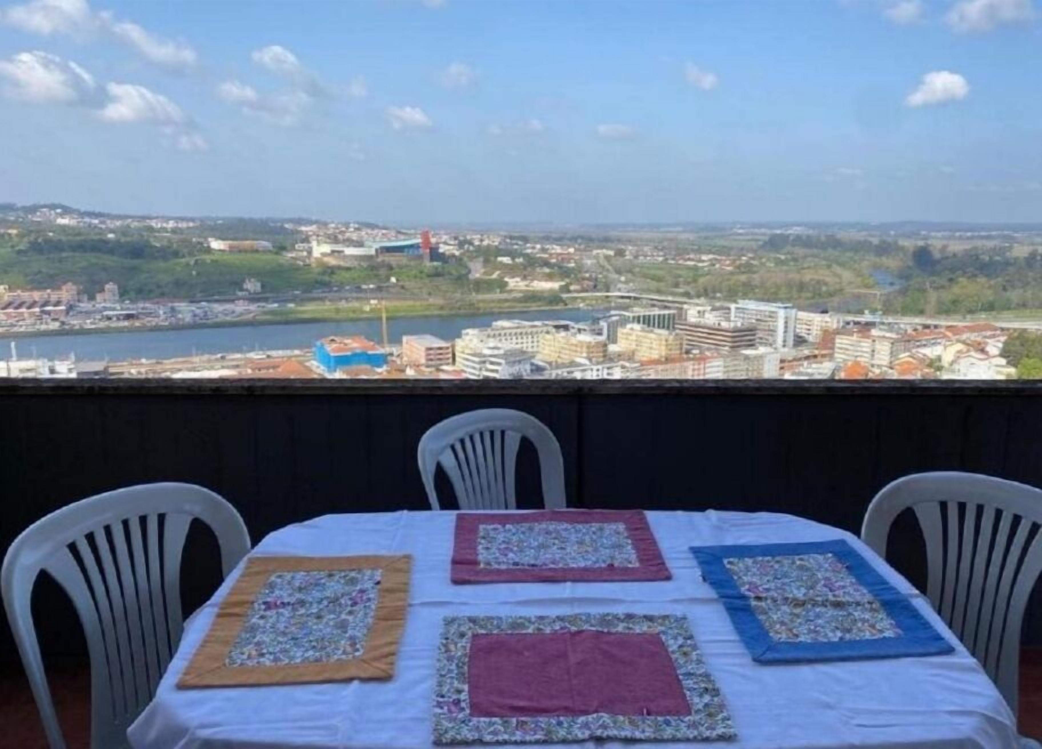 A balcony with a table set for two, overlooking a cityscape with buildings and a river.