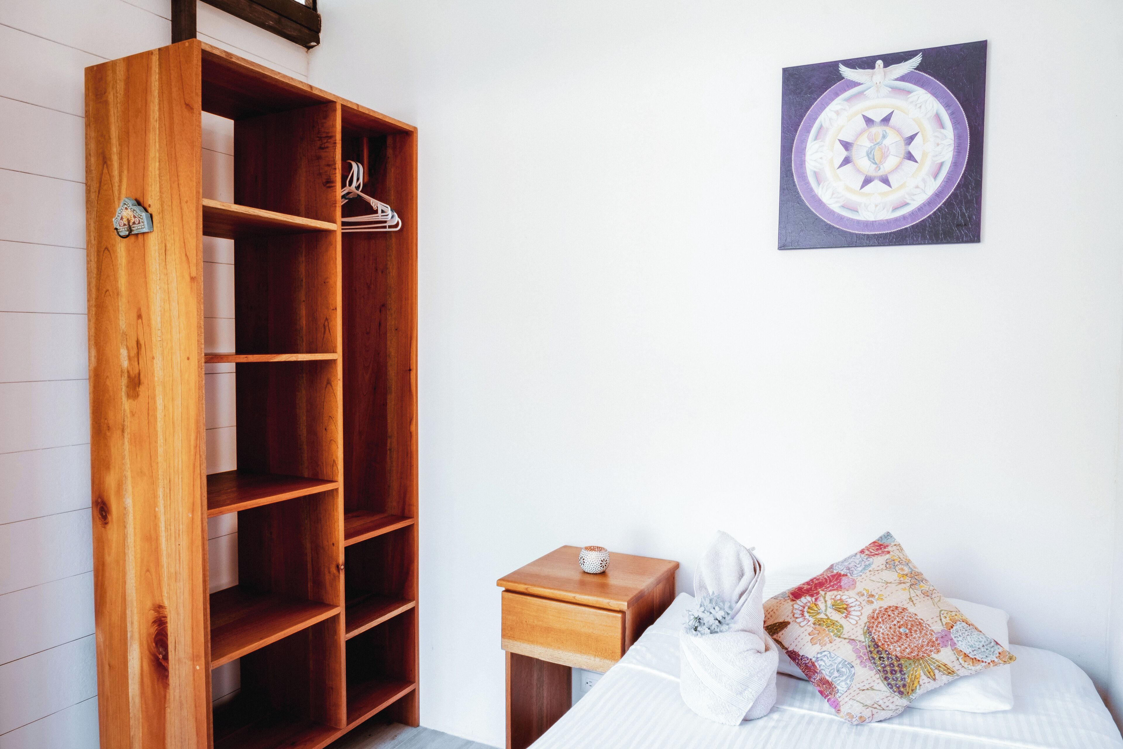 A bedroom with a wooden wardrobe, a small bedside table, a bed with white bedding, and a wall-mounted artwork.