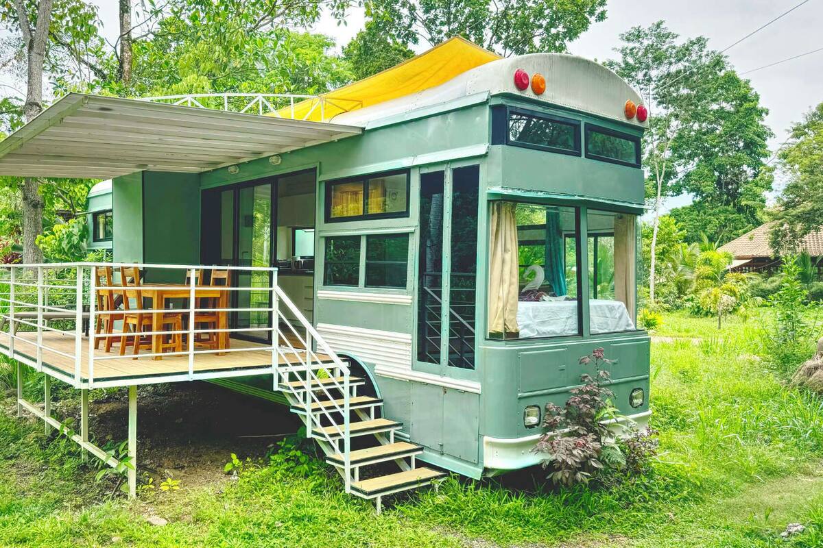 A green bus converted into a house with a wooden deck, stairs, and a small garden area.
