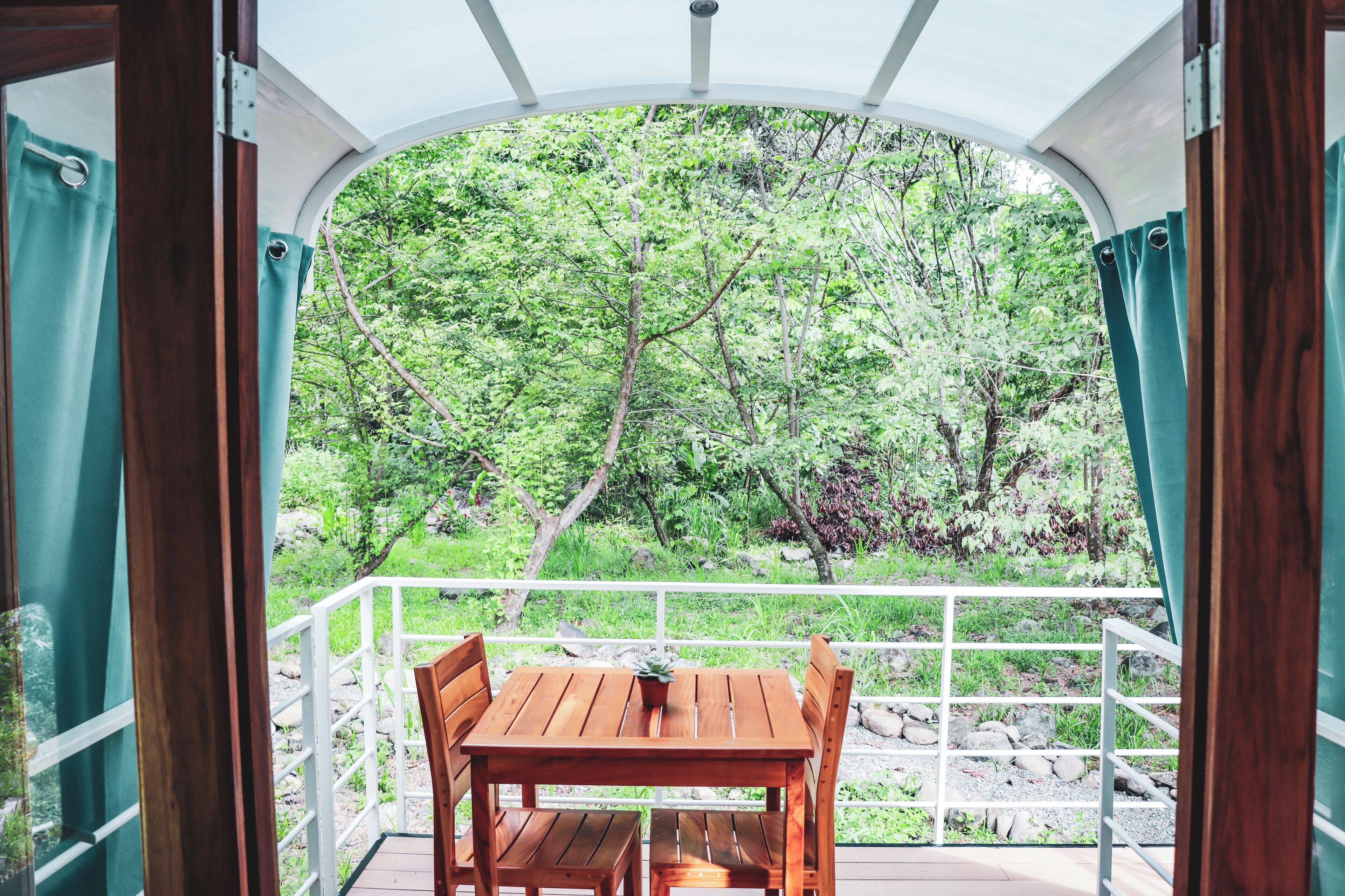 Ein Holztisch mit Stühlen auf einem Balkon mit weißer Brüstung, mit Blick auf eine üppige grüne Landschaft mit Bäumen und Felsen.