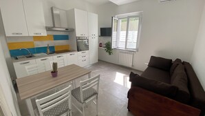 Une cuisine moderne avec des armoires blanches, un four encastré et une fenêtre. On y trouve une table à manger avec des chaises, ainsi qu’un canapé marron dans l’espace salon.