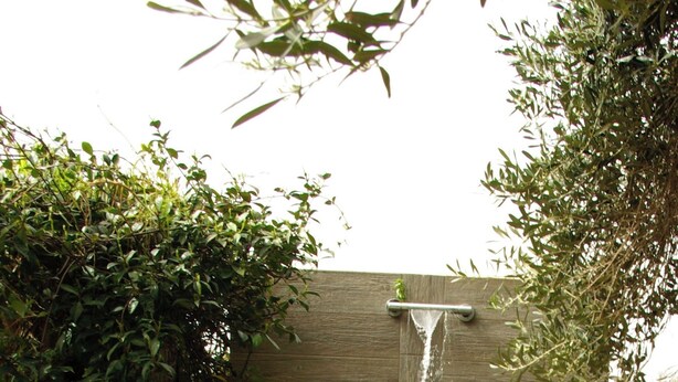 Une fontaine en bois avec une cascade, entourée de verdure et bordée d’un chemin en béton.
