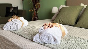 A hotel room with a bed featuring rolled towels and a decorative bow, a green wall, a plant, and a couch with a pillow.