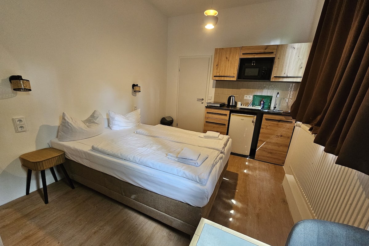 Habitació d'hotel senzilla amb llit, petit tamboret, petita cuina i radiador.