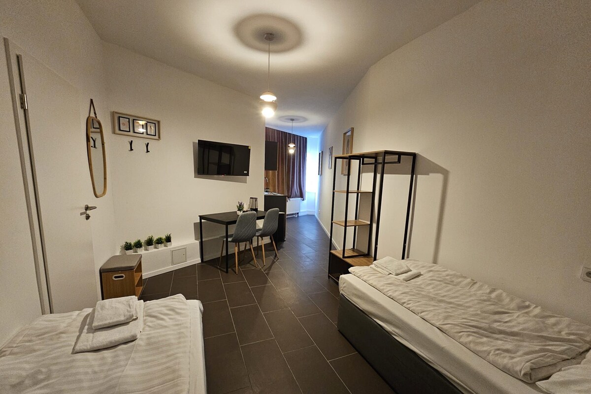 Una habitación de hotel compacta con dos camas individuales, un escritorio, una silla y un televisor montado en la pared.