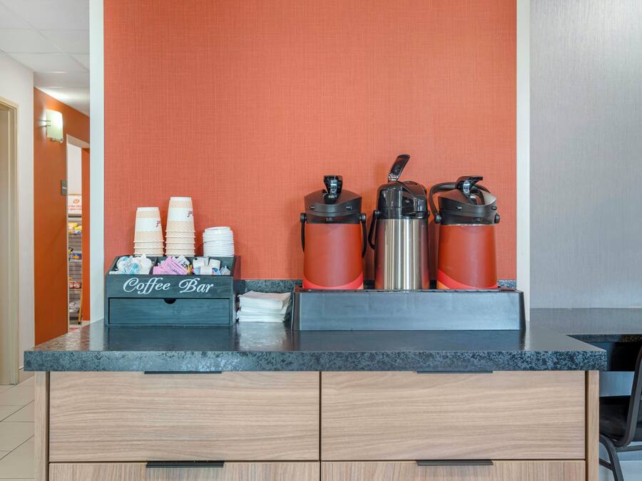 A coffee bar with a coffee maker, a hot water dispenser, and a thermos. There are stacked paper cups and a coffee station sign.
