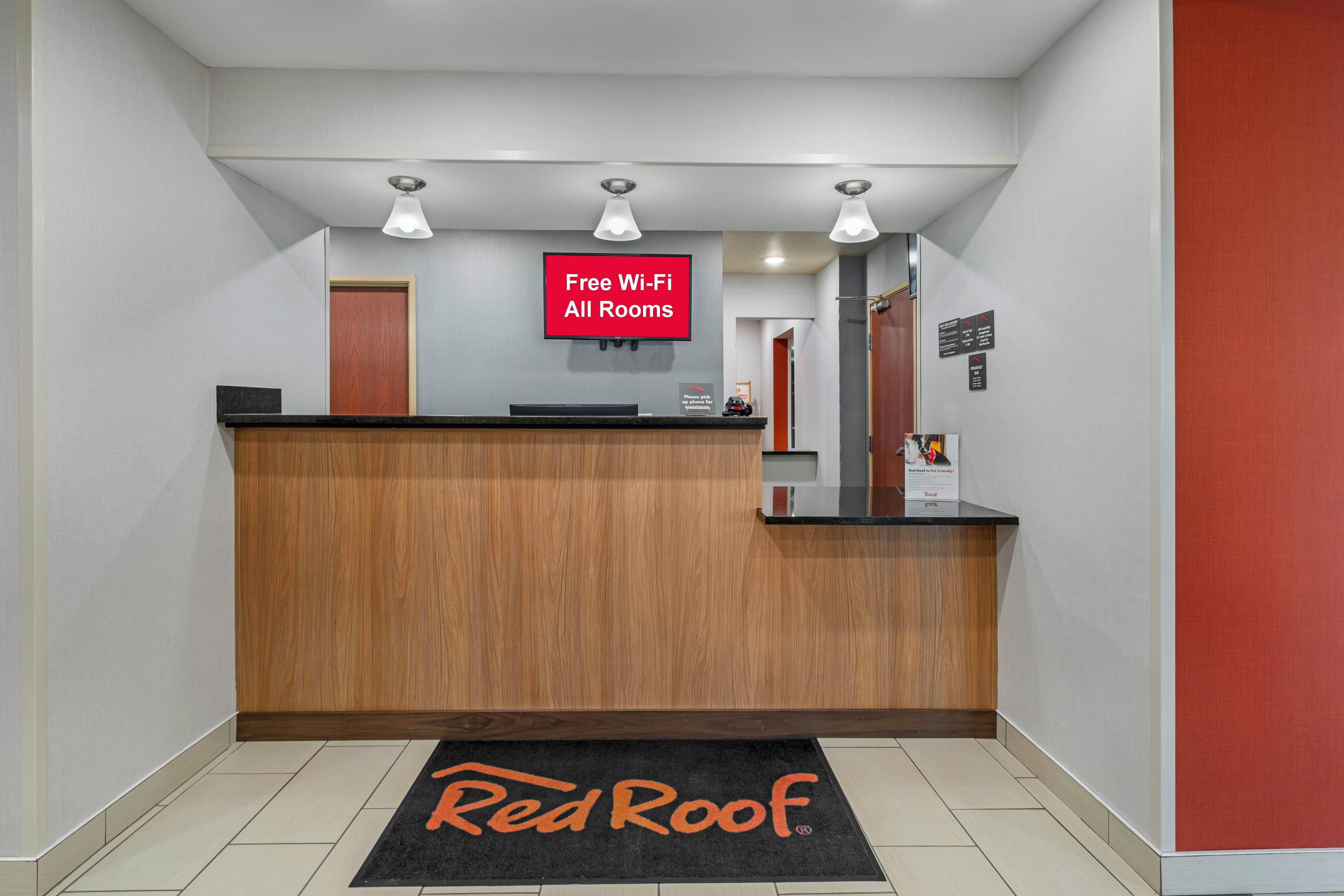 Zona de recepción de hotel con un mostrador de madera, un letrero que indica Wi-Fi gratuito en todas las habitaciones y un felpudo con la marca Red Roof.