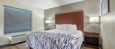 A hotel room with a large bed, two bedside lamps, a wooden headboard, a small nightstand, and a window with blinds.