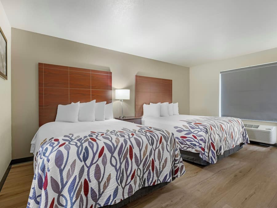 A hotel room with two beds, a nightstand, a lamp, and a window with blinds.