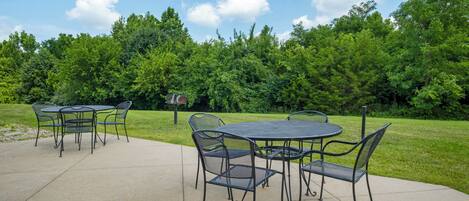 An outdoor patio with metal tables and chairs, surrounded by a grassy area and trees.