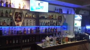 Uma área de bar com prateleiras de garrafas de bebidas, uma tela de televisão e uma máquina de café.