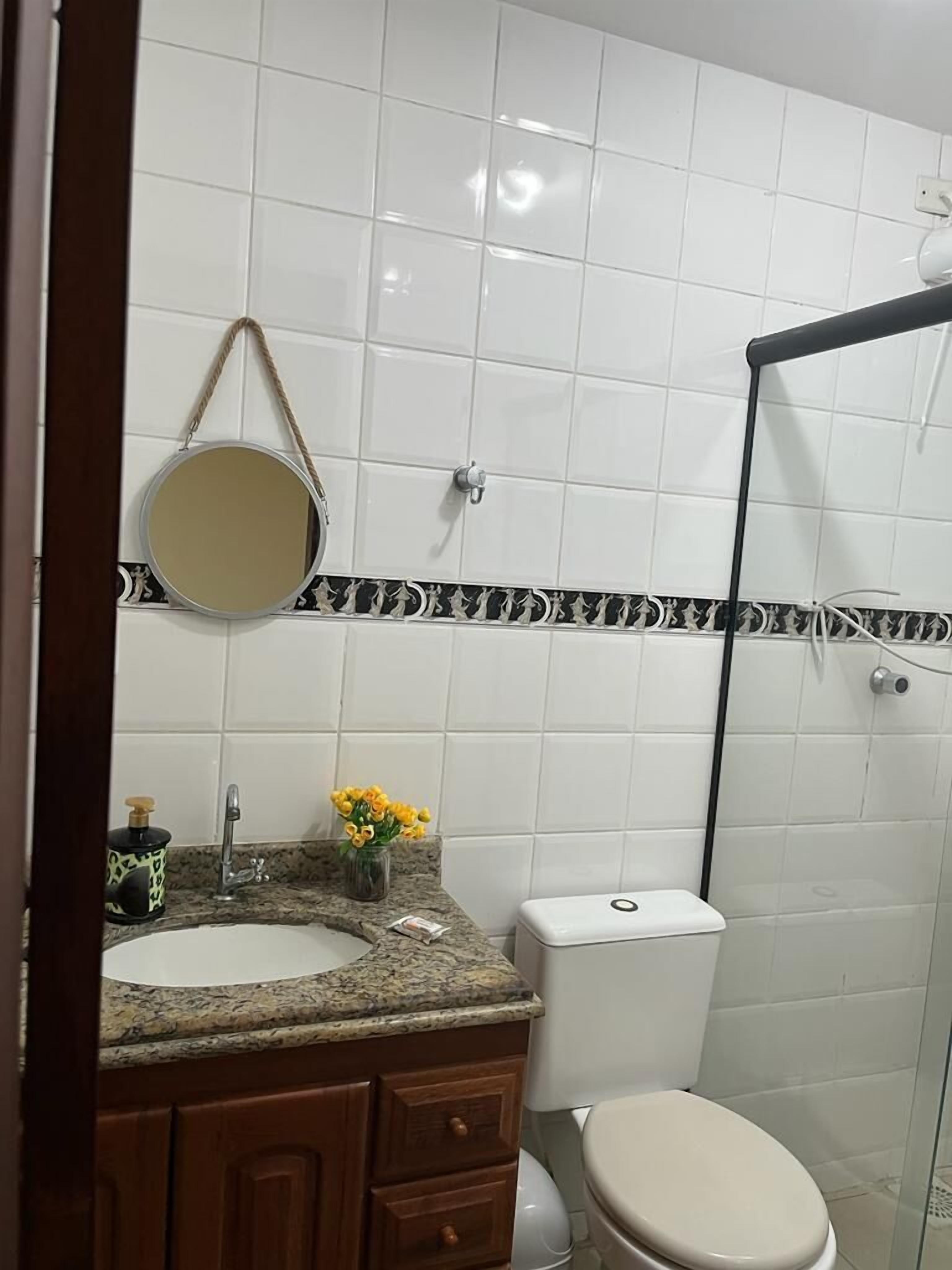 Casa de banho com parede revestida a azulejo branco, espelho redondo, bancada de granito, sanita e cabine de duche.