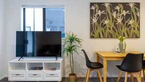 Una sala moderna con un televisor de pantalla plana sobre un mueble blanco, un comedor con una mesa de madera y sillas negras, y un cuadro grande con motivos florales en la pared.