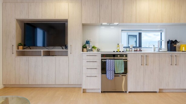 Een moderne keuken met lichte houten kastjes, een ingebouwde oven en een groot raam dat zorgt voor veel daglicht.