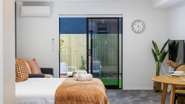 A modern bedroom with a bed, a small table, a chair, and a sliding glass door leading to a balcony.