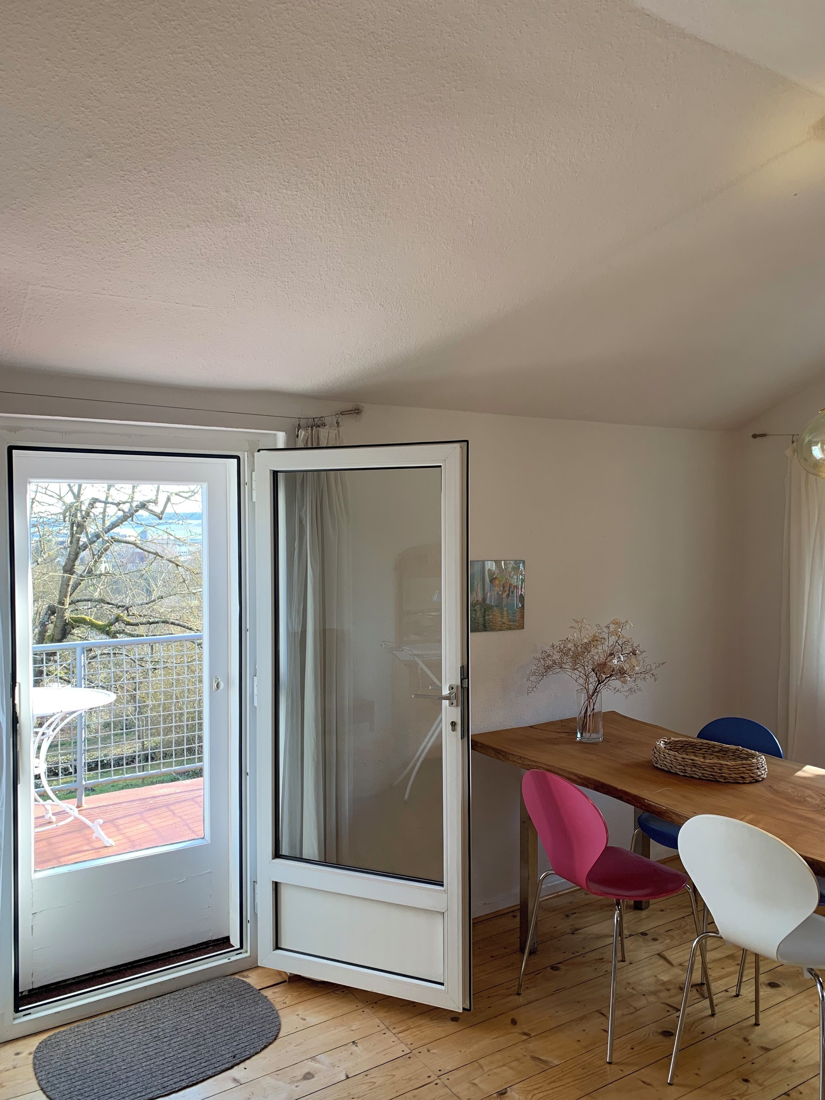 Une pièce comprenant une table à manger en bois, une chaise rose, une chaise blanche et une porte en verre donnant sur un espace extérieur agrémenté d’une clôture en métal et d’un arbre.