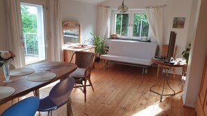 Una habitación acogedora con suelo de madera, una mesa de comedor con sillas, un sofá, un escritorio pequeño con una radio y una ventana con cortinas.