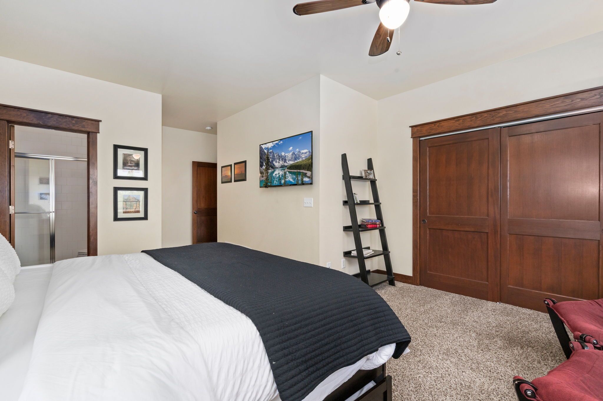 A bedroom with a bed, a ceiling fan, a wardrobe, and a shelf with books.