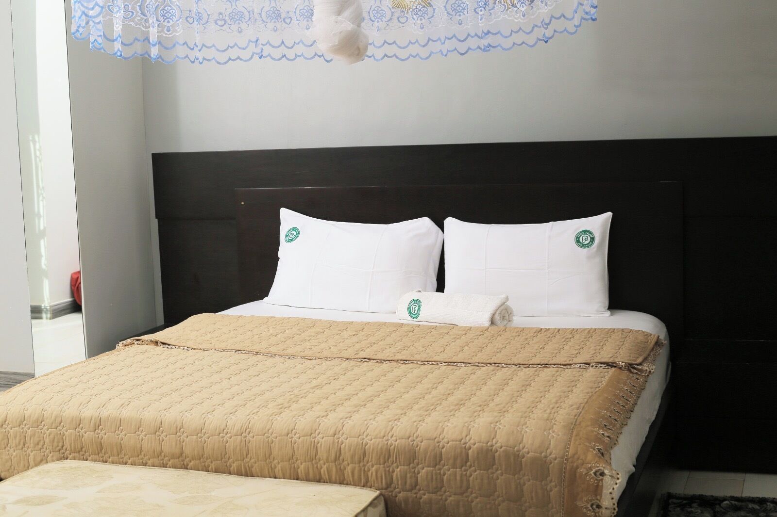 Un lit bien fait, avec une couette beige, deux oreillers blancs ornés d’un logo et une couverture assortie.