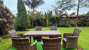 Eine Terrasse mit einem runden Tisch und sechs Korbstühlen, umgeben von üppiger Bepflanzung und einem gepflegten Rasen.