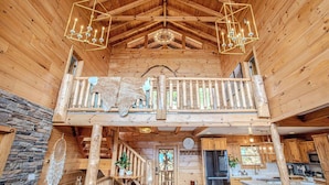 A wooden cabin interior with a balcony, a stone wall, a dreamcatcher, and a kitchen area.