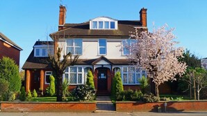 A two-story house with a front porch, a chimney, and a garden with flowering plants. - Ricky Road Guest House (Watford)