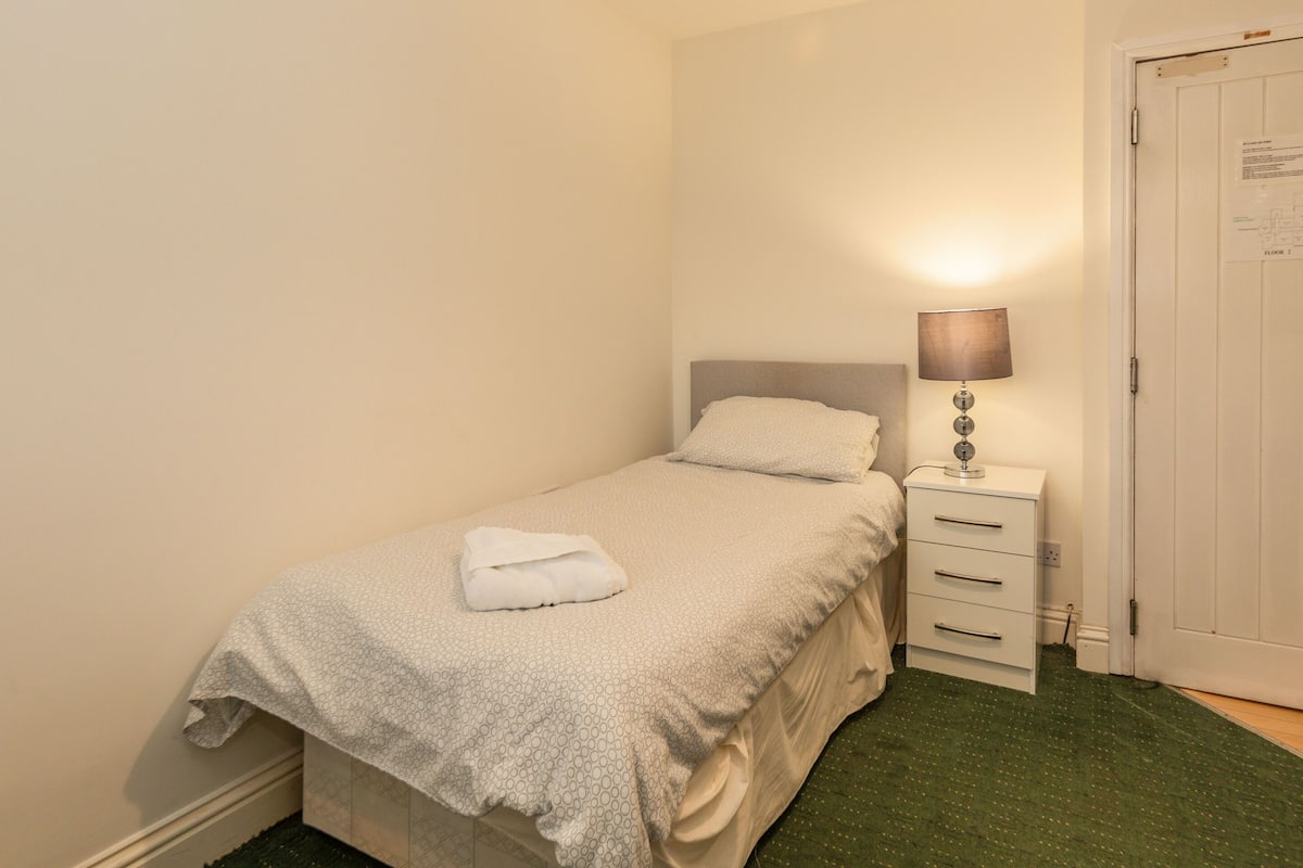 A small, neatly made bed with a white blanket and a folded towel on top, a bedside lamp, a white dresser with drawers, and a closed door.