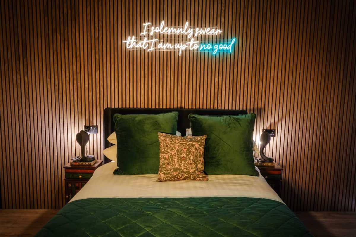 A neatly made bed with a green quilt, two green pillows, and a neon sign above stating 'I solemnly swear that I am up to no good.'