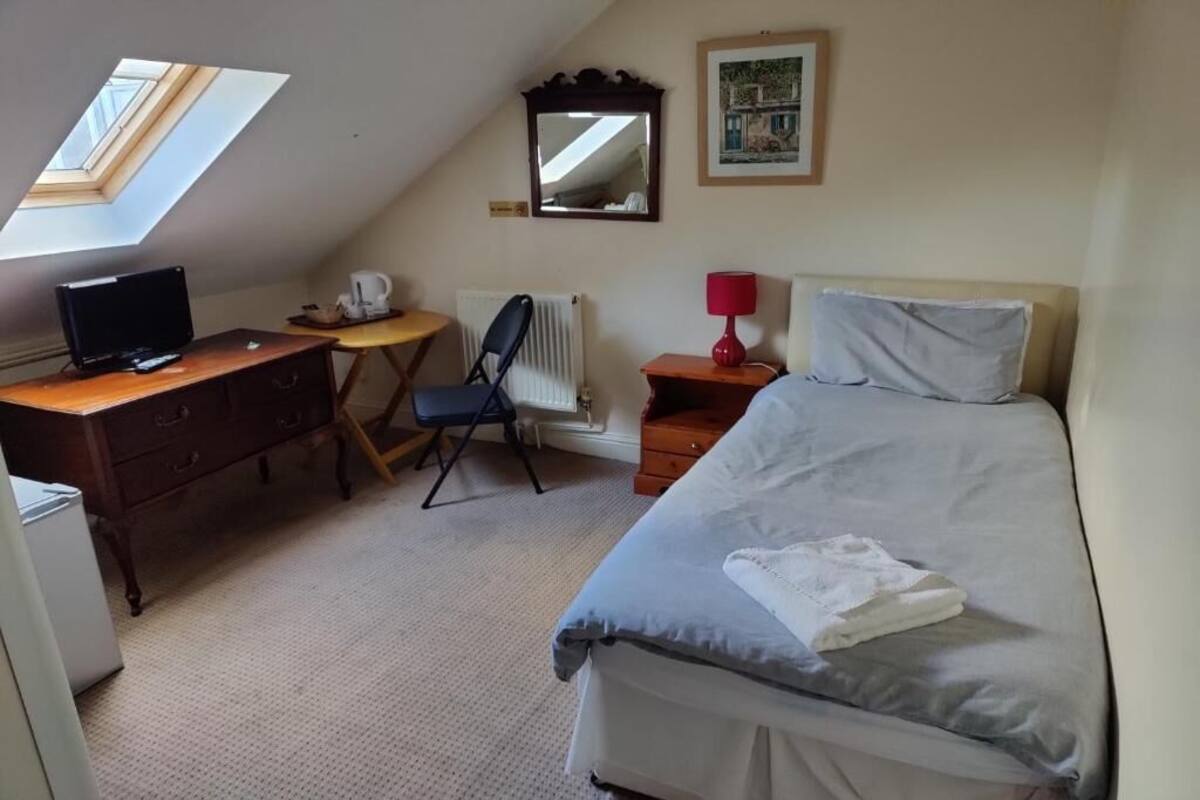 A bedroom with a bed, a desk with a computer, a chair, a radiator, a small table with a lamp, and a window.