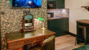 A room with a television mounted on the wall, a desk with drawers, a mirror, a green cabinet, a microwave, and a brick-patterned backsplash. - Ricky Road Guest House (Watford)