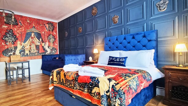 A bedroom with a large bed, a desk, and a tapestry on the wall. - Ricky Road Guest House (Watford)
