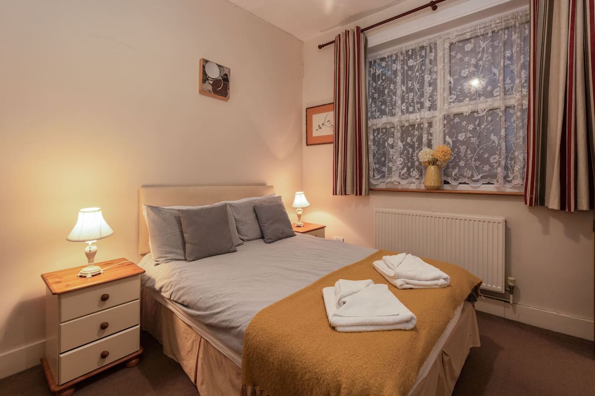 A neatly made bed with a brown blanket, two bedside lamps, a small bedside table with drawers, a window with floral curtains, and a radiator.