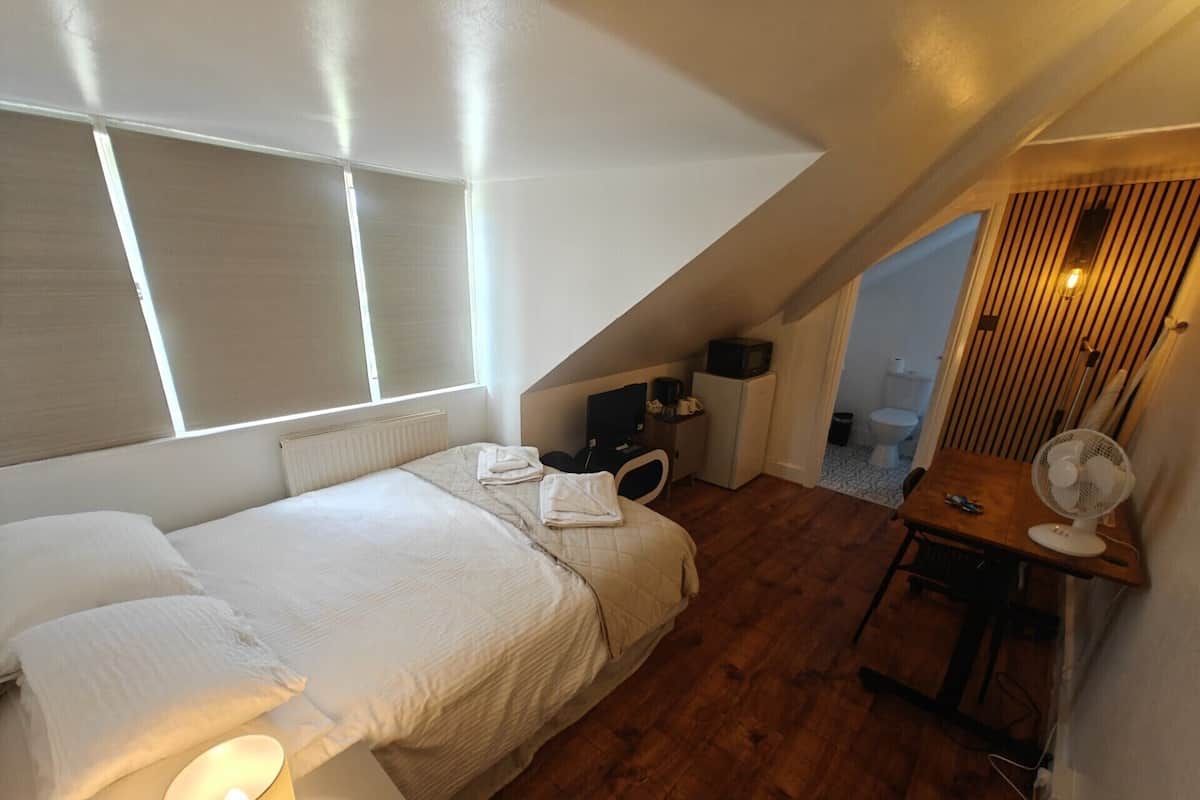 A bedroom with a bed, bedside table, lamp, and a small desk with a fan.