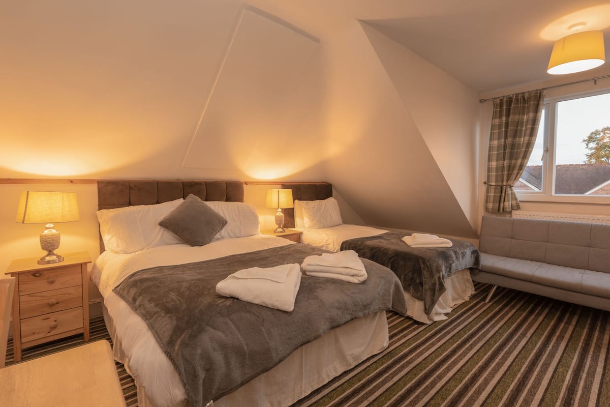 A hotel room with two beds, a sofa, a window with curtains, and a lamp on a bedside table.