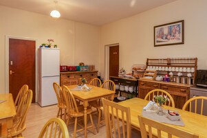Una cocina compacta con refrigerador, armarios de madera, una mesa de comedor con sillas y un estante de pared con vajilla.