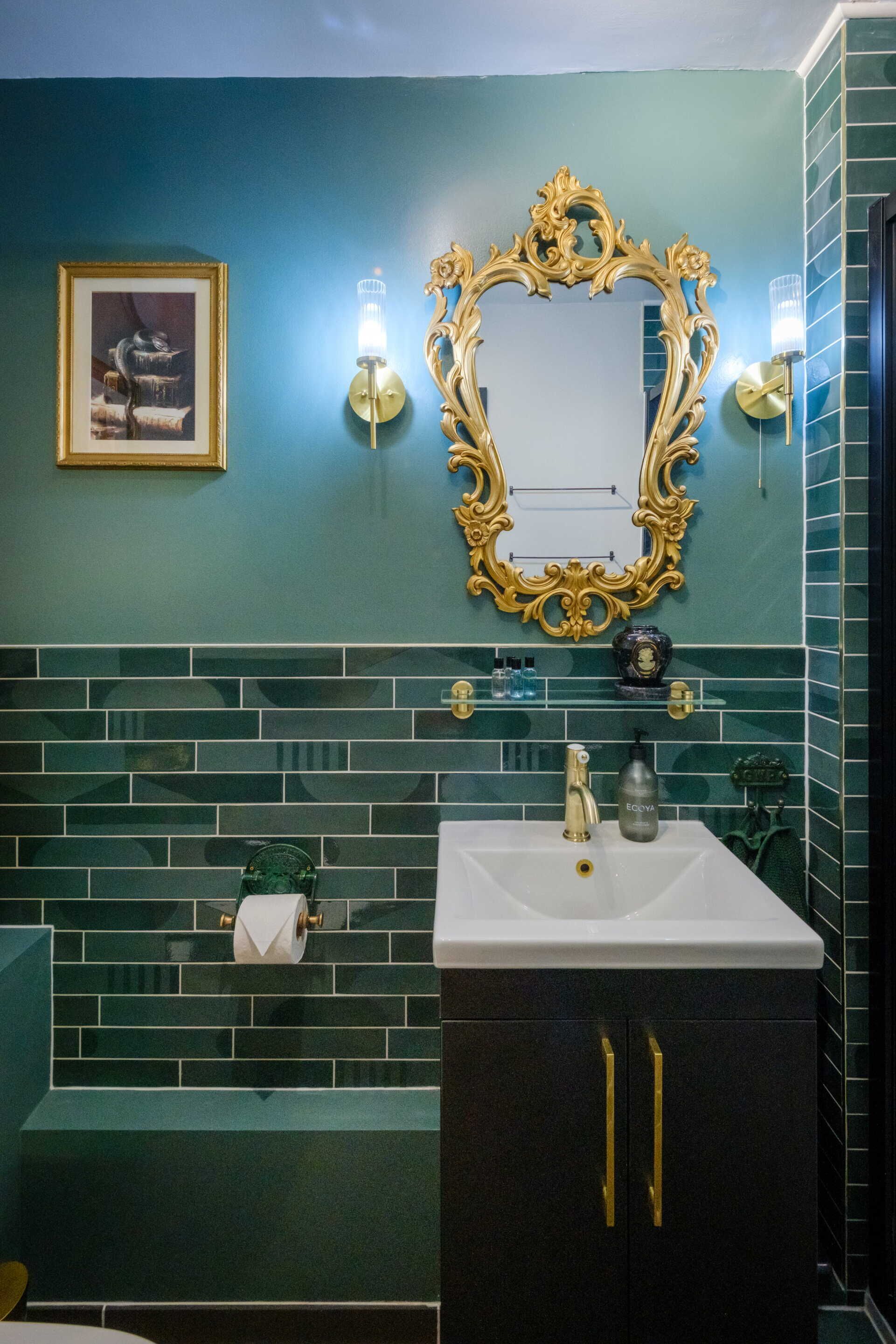 A bathroom with a white sink, gold handles, a gold-framed mirror, and green tiled walls.