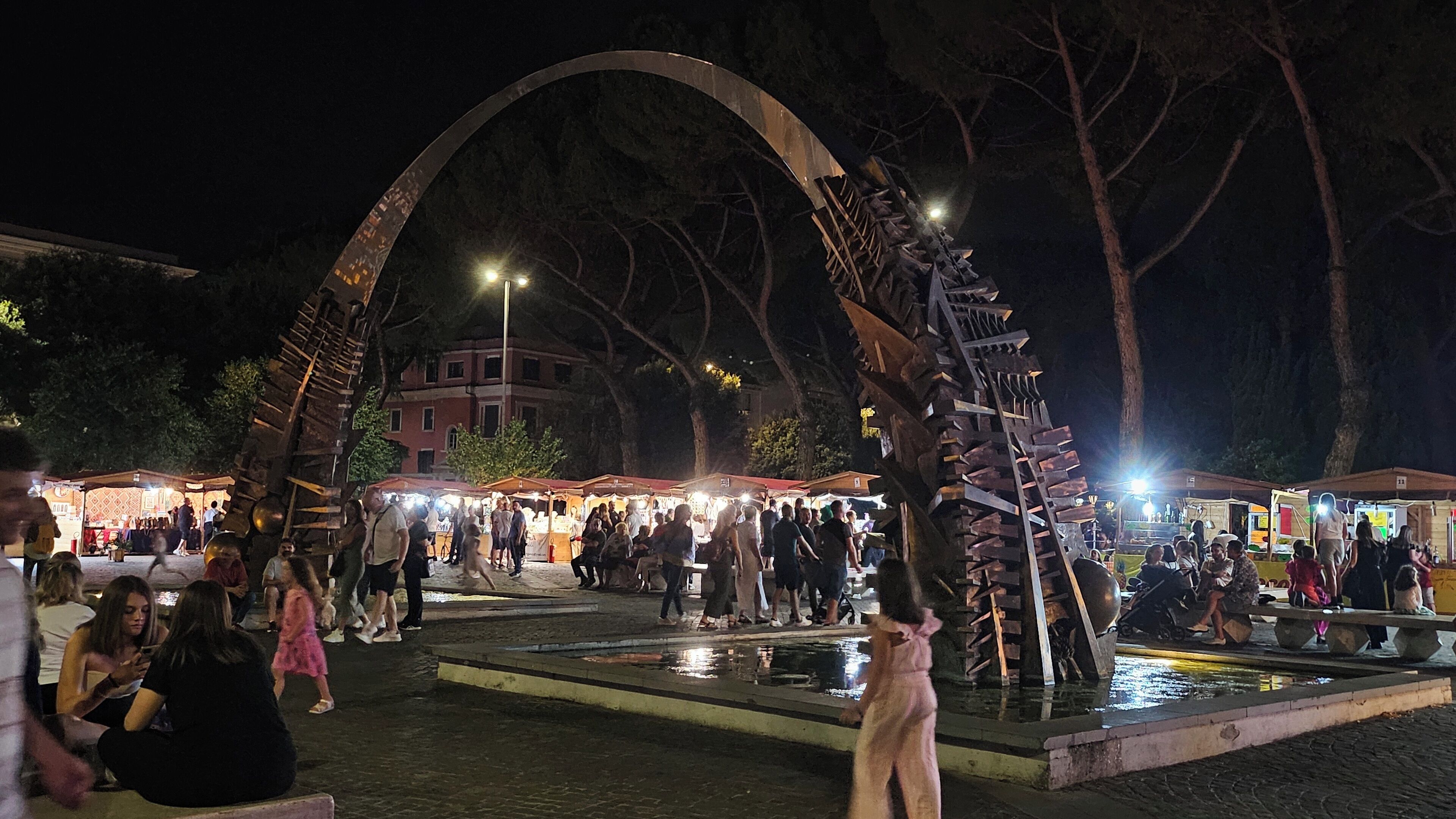 Una piazza pubblica con una grande scultura in metallo, una fontana e bancarelle di mercato.