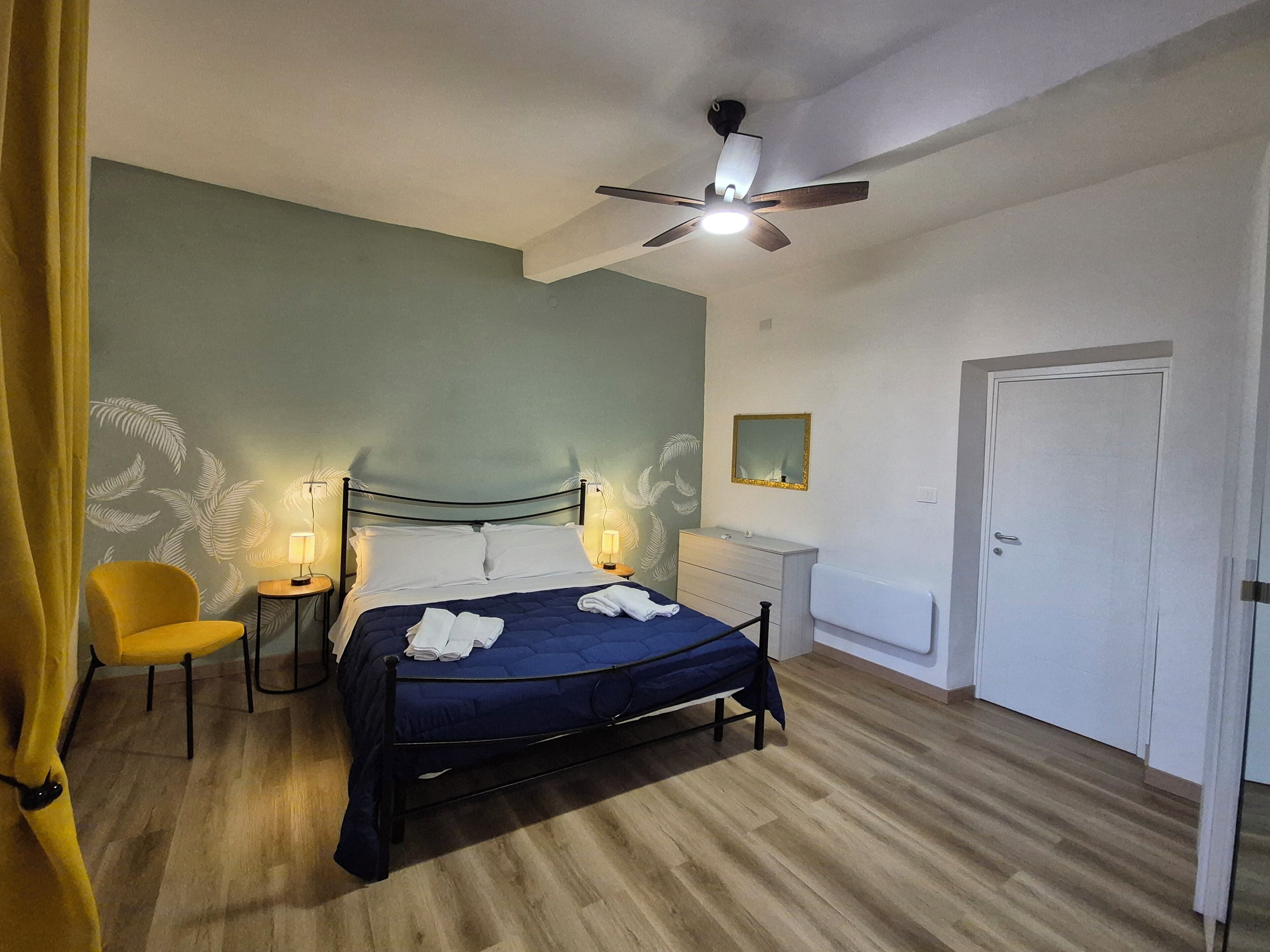 A bedroom with a bed, a ceiling fan, a nightstand, a chair, a dresser, and a door.