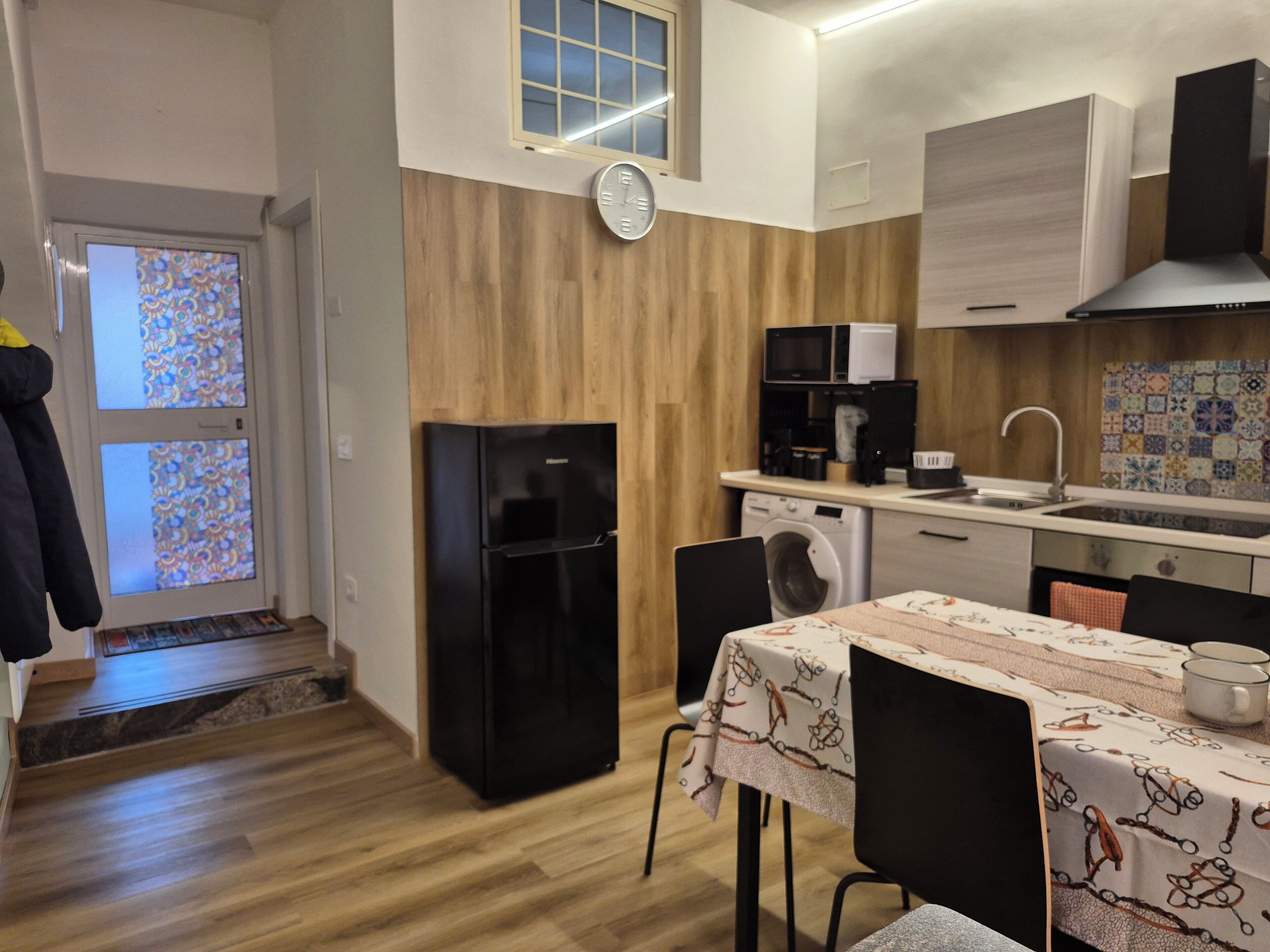 A compact kitchen with a refrigerator, microwave, and washing machine. There's a dining table with chairs, a clock on the wall, and a window with patterned glass.