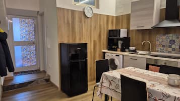 A compact kitchen with a refrigerator, microwave, and washing machine. There's a dining table with chairs, a clock on the wall, and a window with patterned glass.