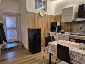 Une cuisine compacte équipée d’un réfrigérateur, d’un four à micro-ondes et d’un lave-linge. On y trouve une table à manger avec des chaises, une horloge au mur et une fenêtre avec du verre décoratif.