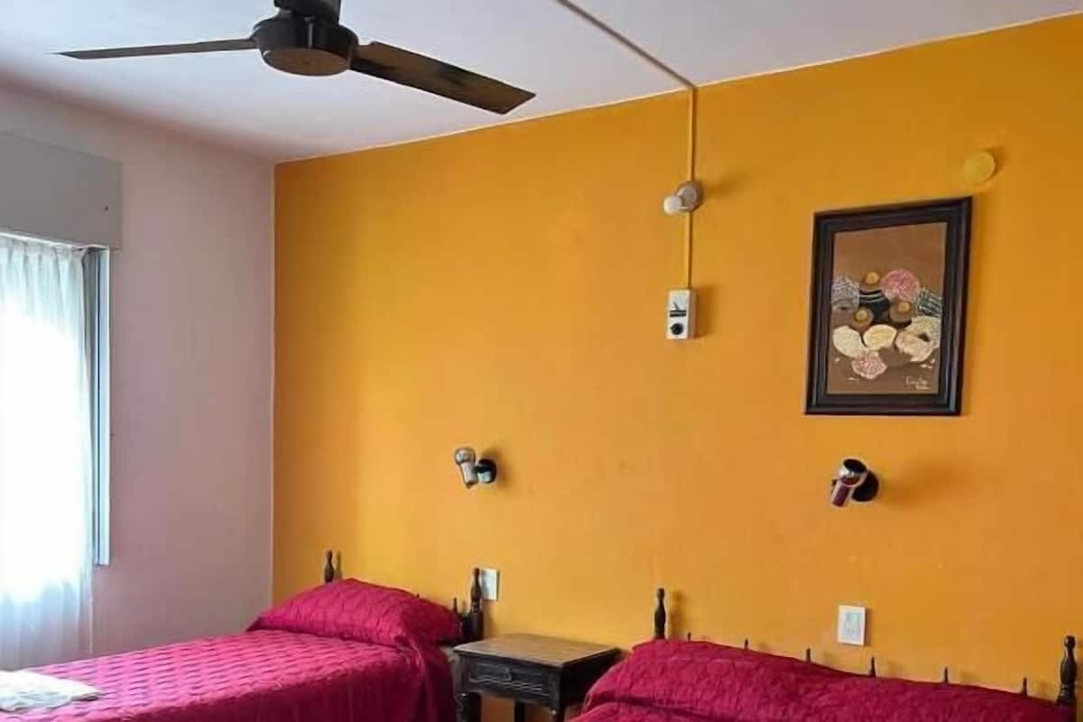 A hotel room with two beds, a ceiling fan, a small table, and a wall-mounted painting.