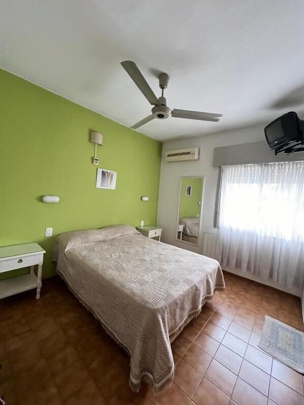 A bedroom with a bed, a nightstand, a ceiling fan, an air conditioning unit, a television mounted on the wall, and a window with curtains.